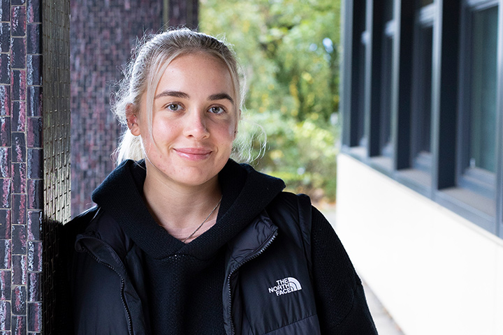 A student leaning against a pillar of a building on campus at the university looking into the camera and smiling