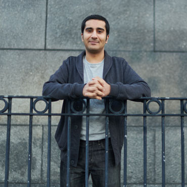 A student standing in front of a stone wall with hands resting on a metal railing and smiling at the camera