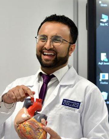 An instructor standing in front of a large digital screen smiling and pointing to a model of a heart