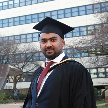 A student wearing a graduation cap and gown stands smiling on campus at the University of Bradford