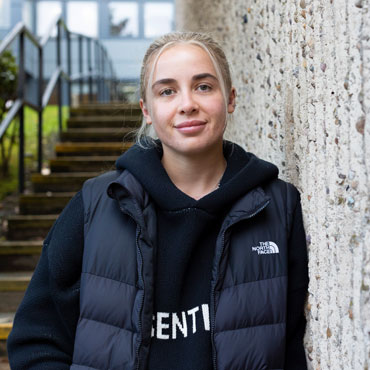 A student is standing outdoors on campus at the University of Bradford smiling at the camera