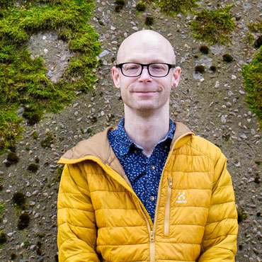 A student smiling at the camera standing in front of a mossy stone wall