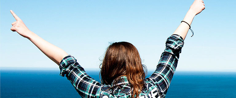 Student with back towards the camera, arms spread, overlooking a sea. The student is wearing a shirt saying 'See the world'.