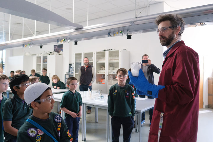 A science technician holding a beaker, speaking to a group of young people