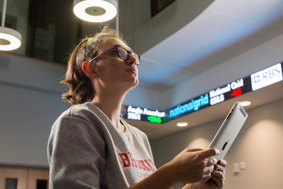 A student holding a tablet computer looking up at the stock ticker at the School of Management