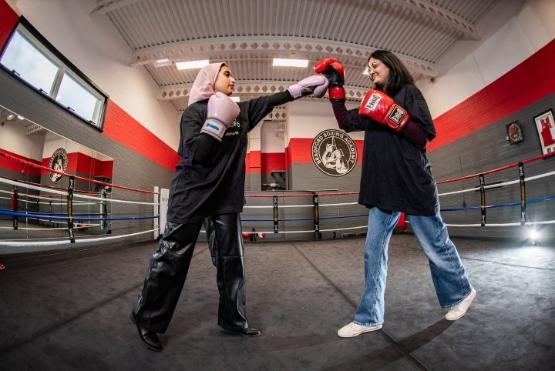 Two students stood up in a boxing ring face each other with one of them pretending to punch the other person