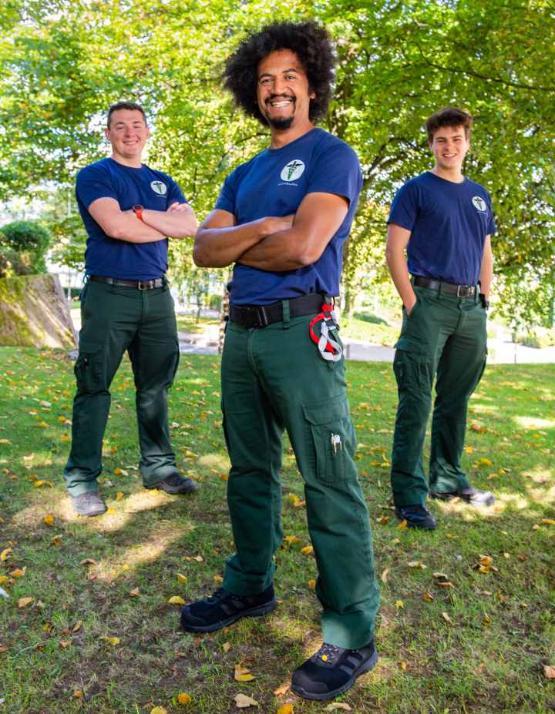 Three people stand in a triangle outside on grassbank wearing T-shirts in sunny weather