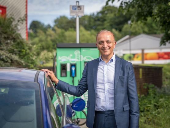 A person dressed in a suit without a tie stands next to a car at at electric vehicle charging point