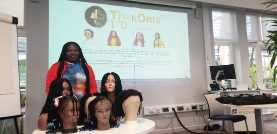 A person stands behind a table which has a number of mannequin heads that are wearing wigs on it in a room with a large projector screen on the back wall