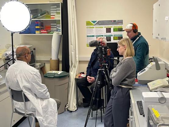 A person sat down dressed in a white lab coat is being interviewed by a three person television crew stood opposite them