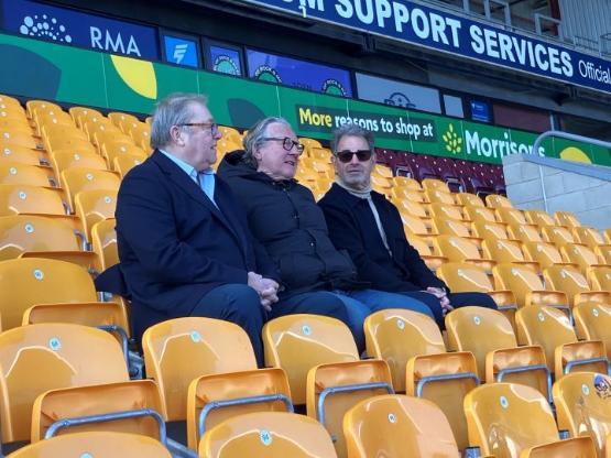 Three people sat next to each other in an otherwise empty football stadium talking to each other