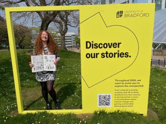 An academic stands holding a closed comic book inside a giant yellow coloured structure
