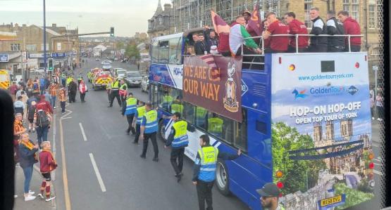 An open top bus filled with people wearing tracksuits on the top deck travels down a street with security guards walking next to it and fans on the pavement