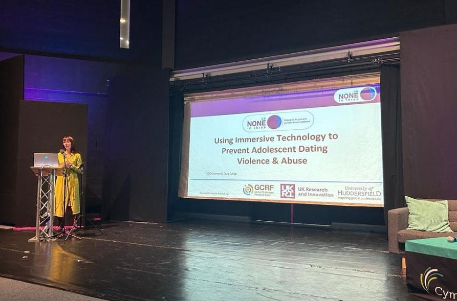 Gill Kirkman standing at a lectern on stage in front of a large presentation screen