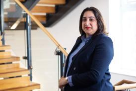 A smartly dressed person facing the camera, leaning on the banister of a staircase in a bright, modern corridor.