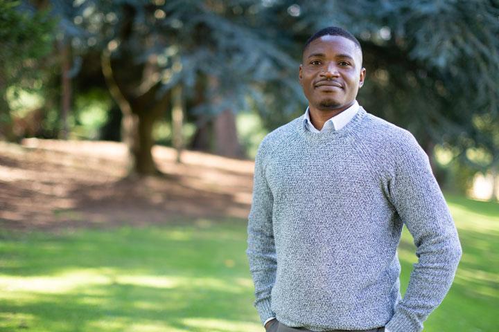 A smartly dressed person standing facing the camera, in a grassy outdoors space with trees in the background.