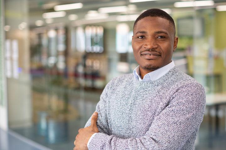 A smartly dressed person standing in a large indoor space, facing the camera with arms folded.