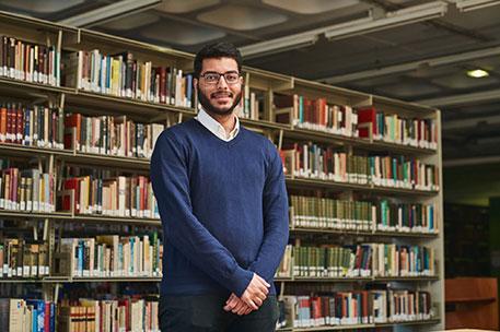 A smartly dressed student standing in front of a book shelf in the library.