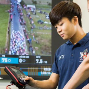 A student and instructor handle a piece of equipment with a large screen showing a sports event in the background.