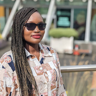 A person standing outside wearing sunglasses. A university building is in the background.